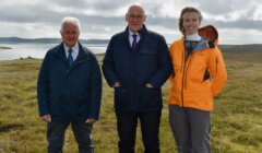Three people stand outdoors on grassy terrain near a body of water under a cloudy sky, all wearing jackets and smiling at the camera.