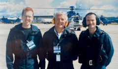 Three men wearing headsets and dark uniforms stand on an airport tarmac with helicopters visible in the background under a cloudy sky.