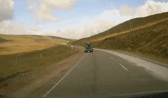A car drives on a rural road through hilly terrain with wind turbines in the distance under a partly cloudy sky.