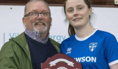 A man and a woman in a blue sports jersey hold a shield-shaped trophy together and smile at the camera.