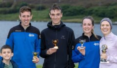 Five people, including a child, stand outdoors by a lake, smiling and holding trophies, with greenery and hills in the background.