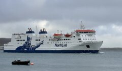A large NorthLink ferry travels on the water under a cloudy sky, with a small boat anchored nearby in the foreground.