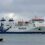 A large NorthLink ferry travels on the water under a cloudy sky, with a small boat anchored nearby in the foreground.