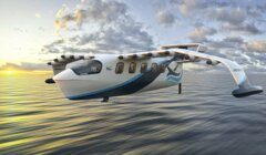 A white and blue electric seaplane with multiple propellers flies low over the ocean at sunset under a partly cloudy sky.