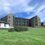 A modern three-story building with large windows and sloped roofs stands on a grassy area, with smaller buildings and bushes nearby under a partly cloudy sky.