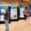 People are discussing information displayed on large presentation boards in a hall with a wooden floor and walls.