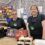 Two people wearing black aprons stand behind a store counter displaying Fox's biscuits. Shelves with snacks and a cash register are visible in the background.