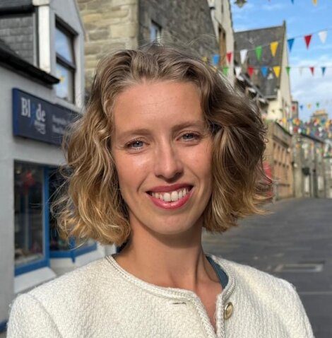 A woman with short, wavy blonde hair and a white jacket stands smiling on a street decorated with colorful bunting and stone buildings.