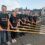 Seven people in matching black shirts stand in a row holding oars, with a boat and buildings visible in the background on a waterfront.