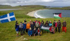 A group of people pose on a grassy hill by the coast, holding Scottish and Palestinian flags, with a beach and sea in the background under a cloudy sky.