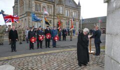 A group of people, some in military uniforms, stand in front of a stone building holding wreaths and flags during a remembrance ceremony. A person reads from a lectern while another stands in silence.