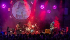 Three musicians perform on stage with drums, guitar, and bass under colorful lighting, with "The Dead Pan" projected behind them. An audience watches from the foreground.