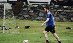 A man wearing a blue vest and black shorts is playing soccer on a grassy field, approaching a white and orange ball with cars and a stone wall in the background.