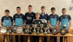 Seven boys in matching sports uniforms stand behind a table displaying numerous trophies and shields in a room with patterned wallpaper.