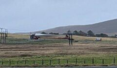 A large wind turbine blade is being transported on a truck through a grassy landscape with hills in the background.