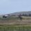 A large wind turbine blade is being transported on a truck through a grassy landscape with hills in the background.