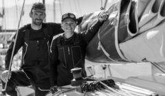 Two people in sailing gear stand on a sailboat, with one resting an arm on a rolled sail; ropes and equipment are visible around them. The image is in black and white.