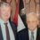 Two men in suits stand side by side in front of a flag with Egyptian colors inside a formal room.