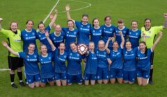 A women's soccer team in blue uniforms poses on a field, smiling and raising their arms, holding a shield-shaped trophy.