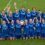 A women's soccer team in blue uniforms poses on a field, smiling and raising their arms, holding a shield-shaped trophy.