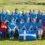 A group of women in blue shirts pose on a golf course with a St. Andrew's Cross (Scottish) flag, some seated and others standing behind.