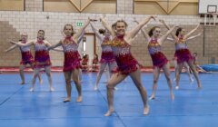 A group of young women in matching sparkly costumes perform a synchronized dance routine on a blue mat in a gymnasium.