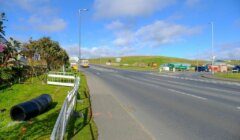 A wide road with multiple lanes runs through a rural area, bordered by grass and some construction materials, under a blue sky with scattered clouds.