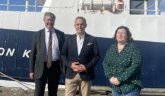Three adults stand side by side on a dock in front of a large white and blue boat. The two men wear suits, and the woman wears a green patterned top.