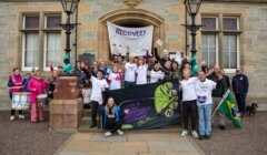 A group of people stand in front of a building holding banners that read "RECOVERY" and "Lived Experience Recovery Organisation." Some are wearing matching T-shirts and appear to be celebrating.