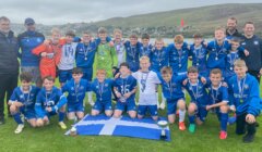 A youth soccer team in blue uniforms poses outdoors with coaches behind a Scottish flag and trophies. All are wearing medals and smiling. Hills and cloudy sky are visible in the background.
