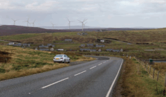 A white car is parked on the side of a rural road with scattered houses and wind turbines visible on rolling hills in the background.