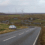 A white car is parked on the side of a rural road with scattered houses and wind turbines visible on rolling hills in the background.