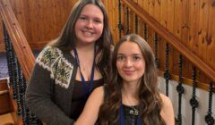 Two young women with lanyards pose indoors by a wooden staircase; one is standing, and the other is seated.