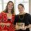Two women stand indoors, one in a red dress and one in black, smiling and holding a plaque reading "Scots Language Awards, Scots Writer of the Year.