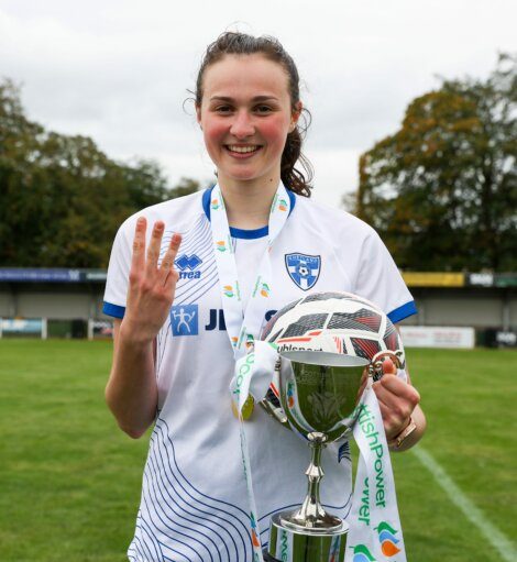 A smiling female soccer player in a white and blue jersey holds a trophy and a soccer ball, showing three fingers, with a soccer field in the background.