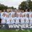 A women's soccer team poses on a field behind a "WINNERS" sign with medals around their necks and a trophy in front of them. Coaches and staff stand on either side.