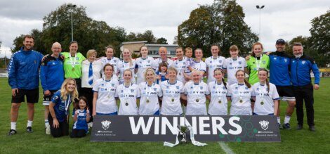 A women's soccer team poses on a field behind a "WINNERS" sign with medals around their necks and a trophy in front of them. Coaches and staff stand on either side.