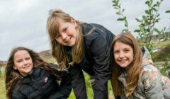 Three girls in jackets smile outdoors on a cloudy day, with greenery and a small tree in the background.