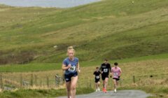 A woman in athletic attire runs uphill on a rural road, followed by four other runners, with grassy hills and water in the background.