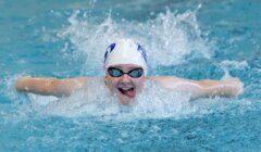 A swimmer wearing goggles and a swim cap performs the butterfly stroke in a pool, creating a splash in the water.
