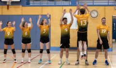Six volleyball players in yellow and black uniforms stand in a gym; five raise their arms while two teammates high-five, and one stands relaxed to the right.