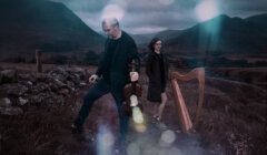 Two people stand outdoors in a hilly landscape; one holds a violin, the other stands by a harp. The scene is dark with lens flares and mountains in the background.