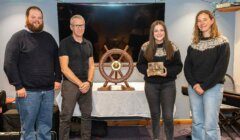 Four people stand indoors in front of a ship’s wheel display; one person holds a plaque or award and they are all facing the camera and smiling.