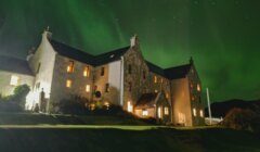 A large, old stone building with lit windows at night, under a green sky illuminated by the northern lights.