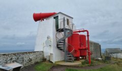A white lighthouse building with a spiral metal staircase, large red foghorn, and red tanks, set near a stone wall overlooking the sea under a cloudy sky.