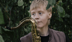 Person with short blonde hair holding a brass saxophone stands among green leaves, looking slightly to the side.