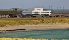 A small airport terminal building labeled "Sumburgh Airport" stands near a beach with grassy dunes and clear blue water in the foreground.