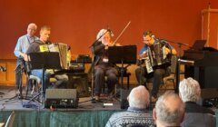 Five musicians perform on stage with accordions, a violin, and a keyboard in front of an audience, against a plain orange wall.