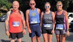 Four people in athletic gear with race bibs stand outside in sunny weather, posing for a photo before a running event.