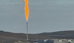A large industrial gas flare burns with black smoke rising above a facility on a grassy, hilly landscape under a cloudy sky.